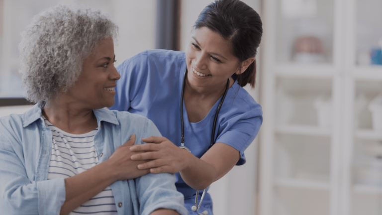 A nurse in blue scrubs smiles warmly at an elderly woman with curly gray hair, placing a supportive hand on her shoulder in a friendly, caring manner.