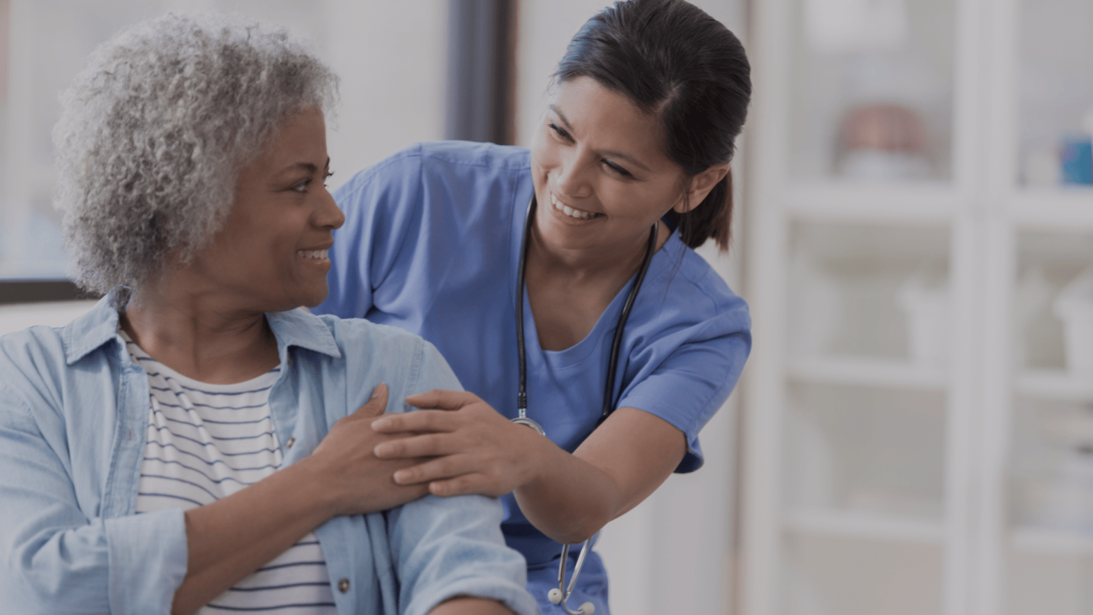 A nurse in blue scrubs smiles warmly at an elderly woman with curly gray hair, placing a supportive hand on her shoulder in a friendly, caring manner.