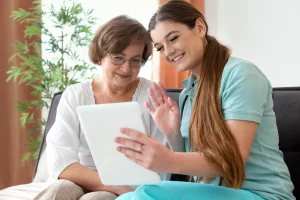 Two women sit on a couch, one older and one younger, smiling and waving at a tablet during a video call.