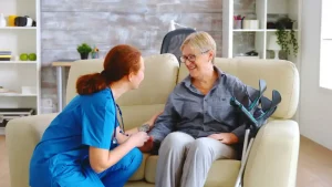 A nurse in blue scrubs smiles warmly at an elderly woman sitting on a beige sofa, holding crutches. They are in a cozy living room setting.