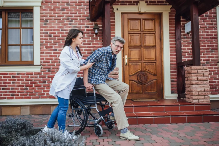 A man pushes a wheelchair while a woman stands beside it, both engaged in conversation outdoors.