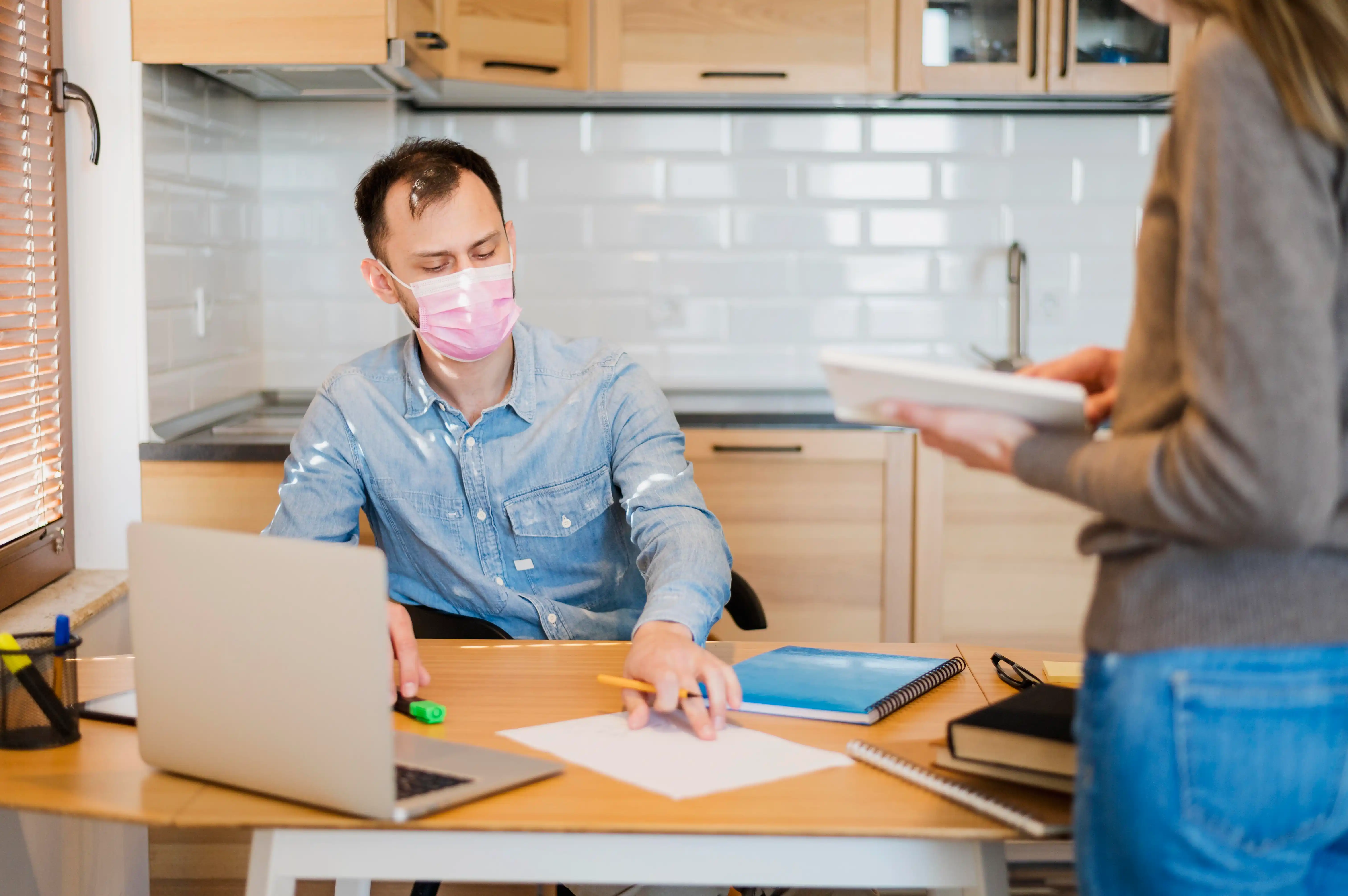 A man and woman wearing face masks sit at a desk, engaged in conversation or working on a task together.
