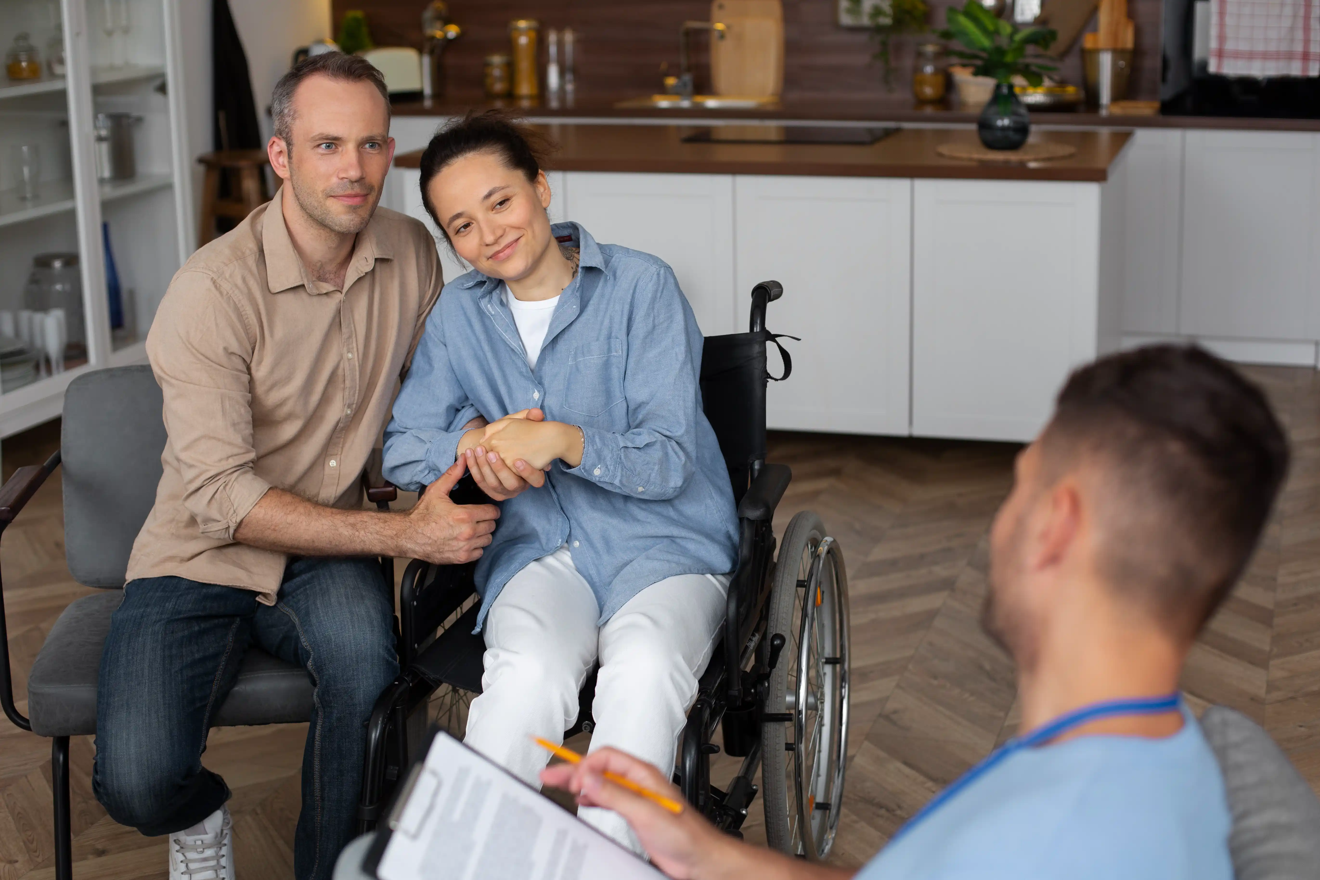 A man and woman in wheelchairs are seated together in a cozy living room, surrounded by warm lighting and furniture.