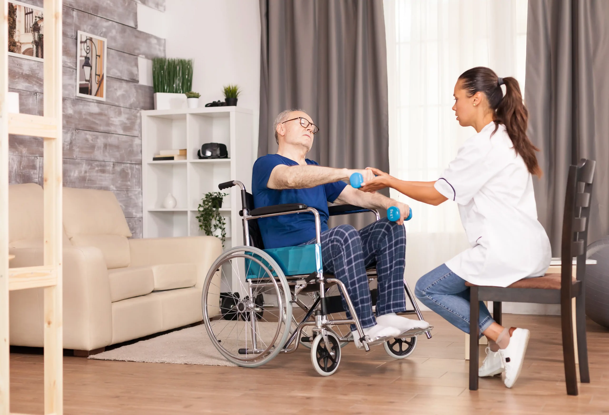 A caregiver assists a senior man in a wheelchair with arm exercises using dumbbells in a cozy living room. The scene conveys encouragement and support.
