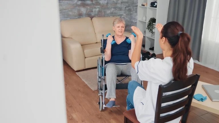 A woman in a wheelchair exercises with small blue dumbbells at home, assisted by a caregiver. The scene is warm and supportive, set in a cozy living room.