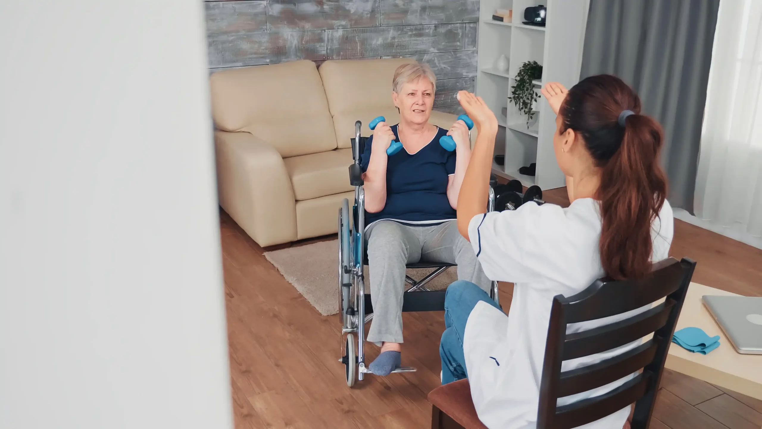 A woman in a wheelchair exercises with small blue dumbbells at home, assisted by a caregiver. The scene is warm and supportive, set in a cozy living room.