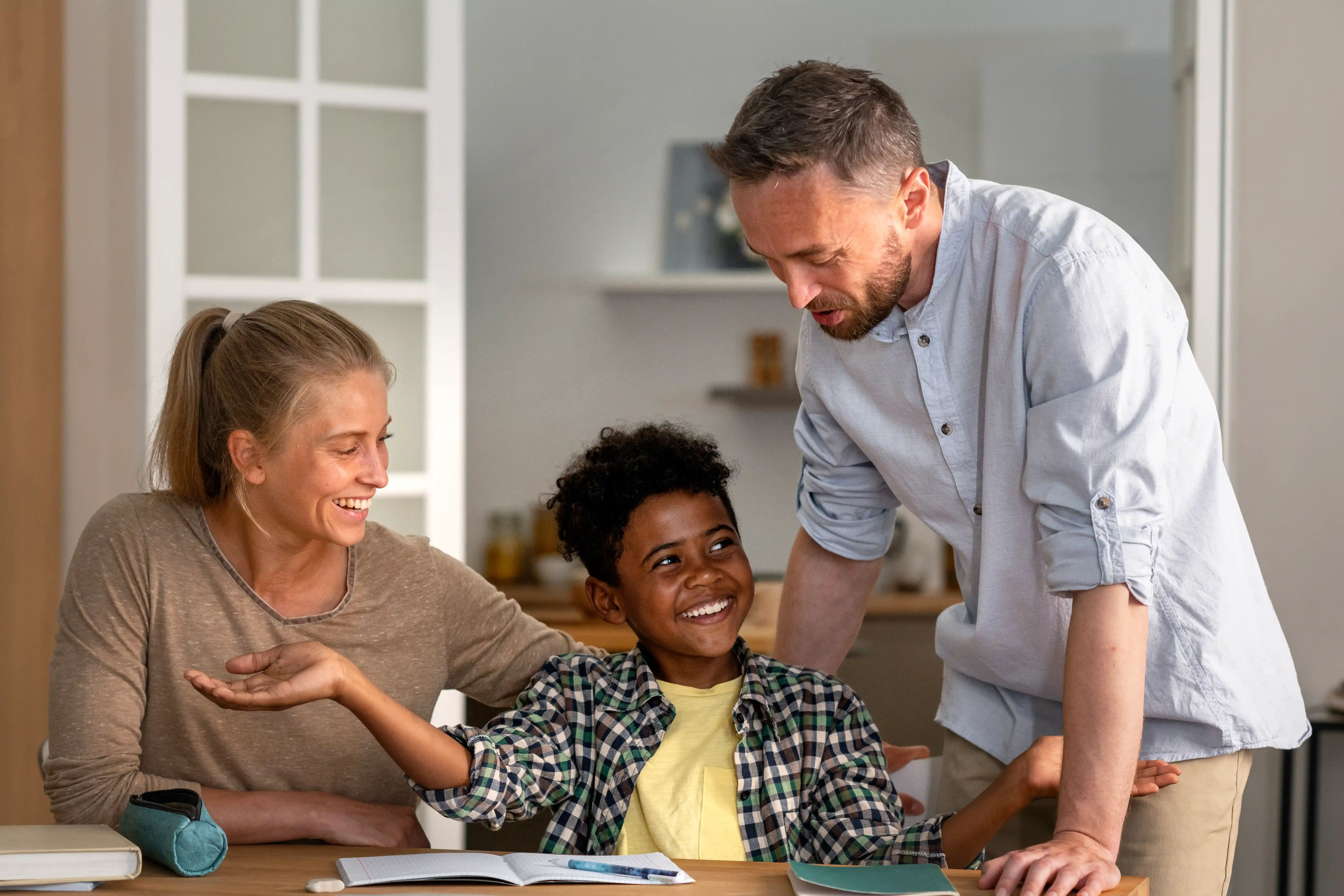 A joyful family studying together at a table, with a child smiling between two adults. The atmosphere is warm and supportive, indicating teamwork.