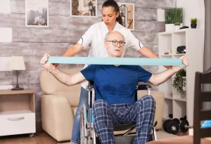 A caregiver assists an elderly man in a wheelchair by helping him stretch a resistance band. The room is cozy, with wooden walls and homey decor.