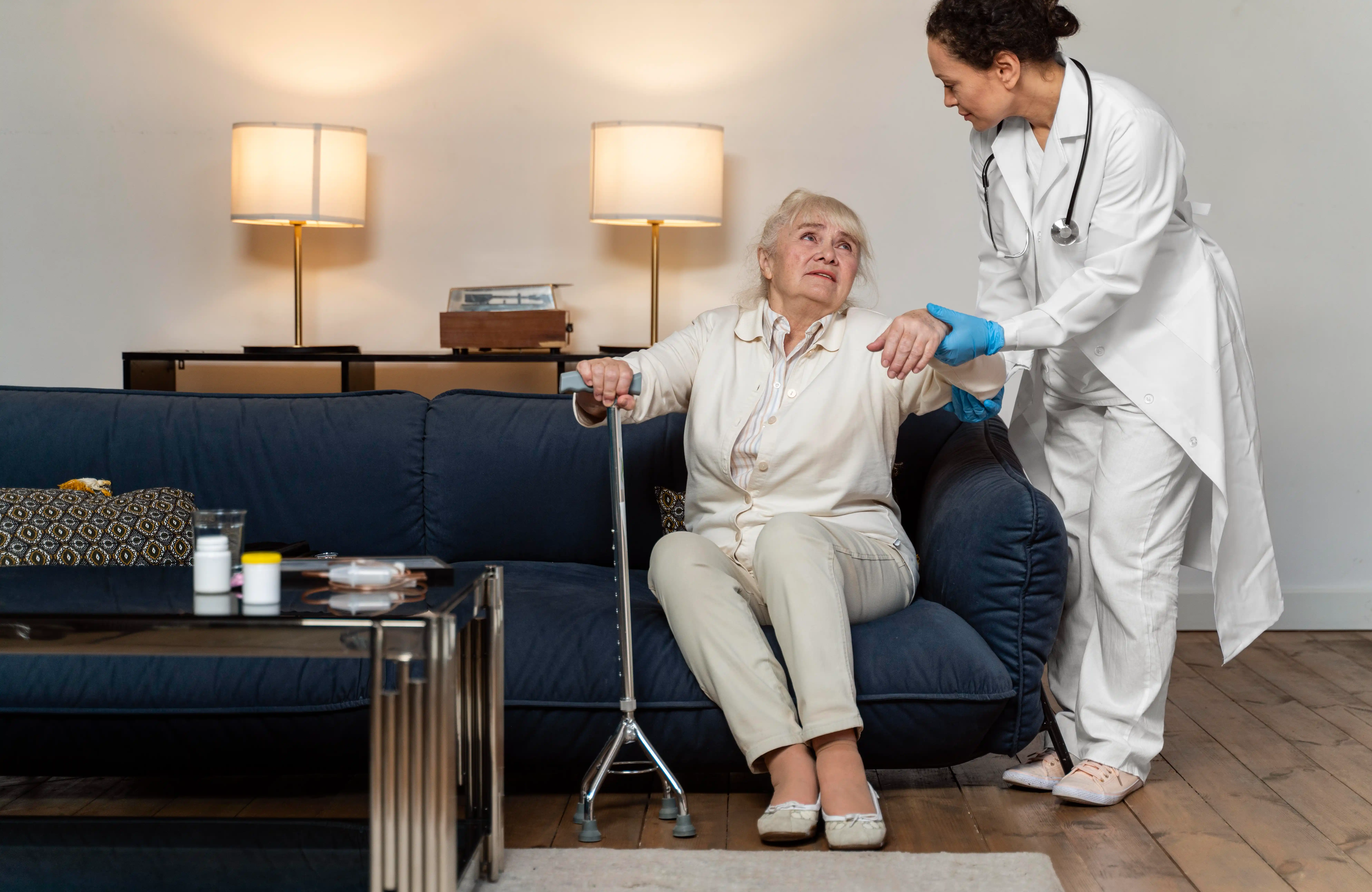 A doctor assists an elderly woman with a cane from a blue sofa in a warmly lit living room. The scene conveys care and support.