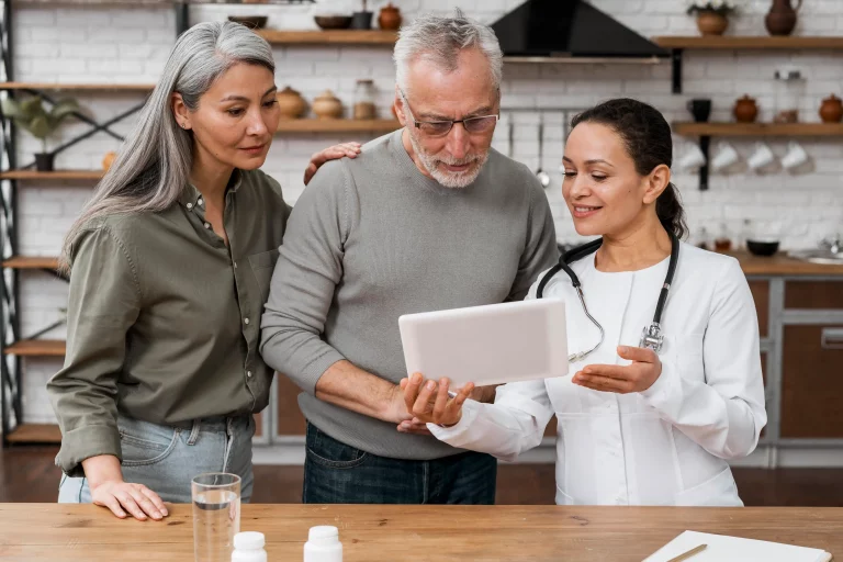 A woman in a lab coat discusses information from a tablet with two individuals in a kitchen setting, surrounded by ingredients.
